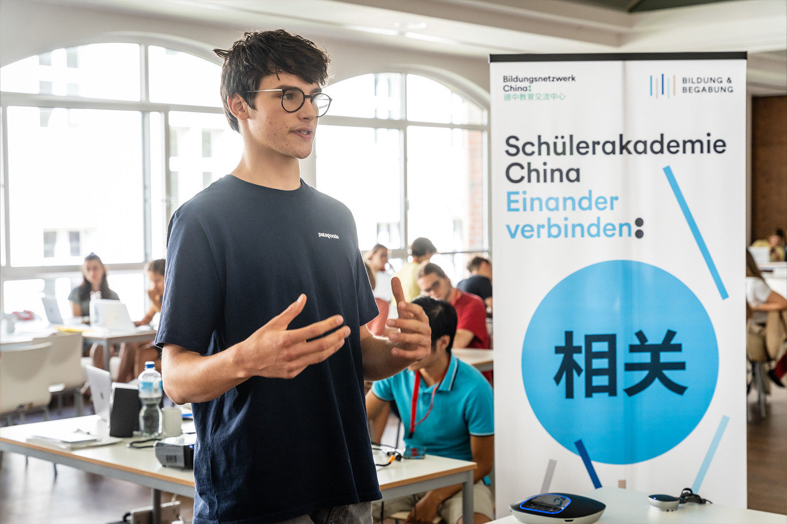 Ein junger Mann mit Brille steht gestikulierend vor einem Aufsteller mit dem Logo der Schülerakademie China. Im Hintergrund sieht man andere Jugendliche. 