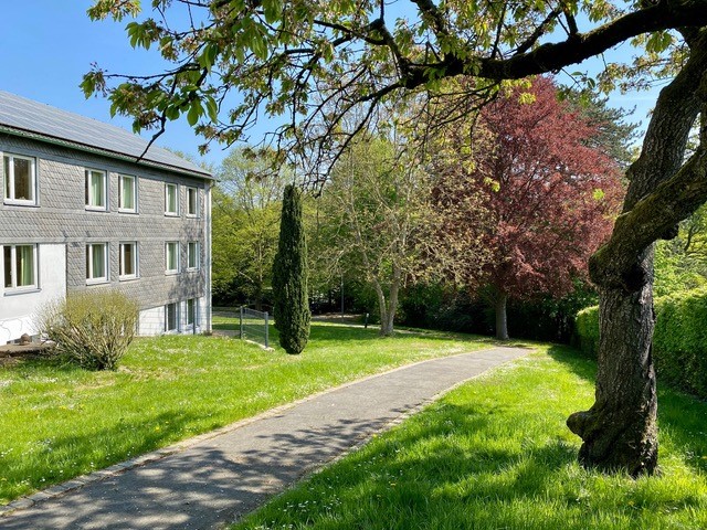 Ein Weg führt bergab an einem Gebäude entlang durch eine Parkanlage.