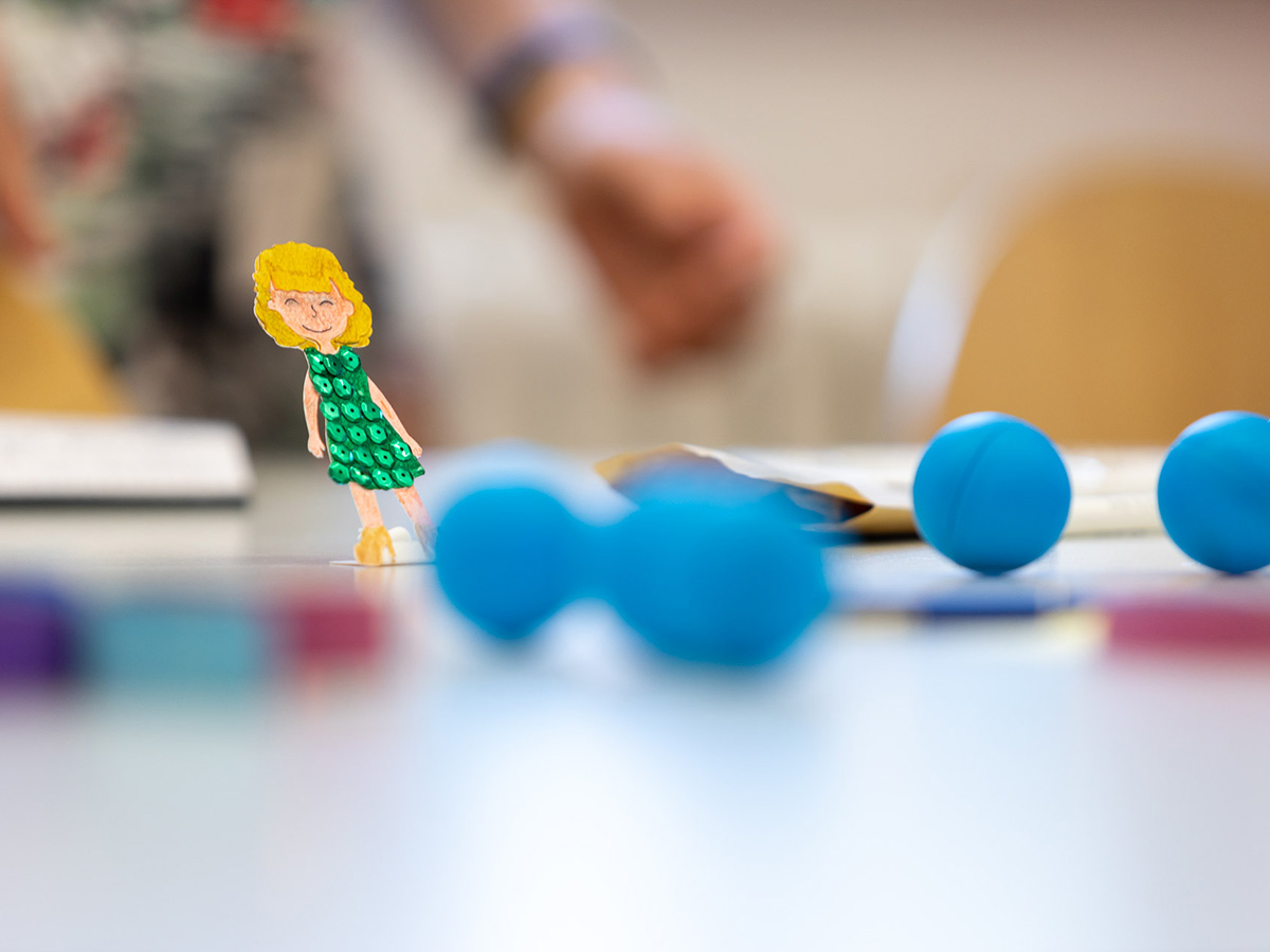 Auf einem weißen Tisch liegen mehrere blaue Kugeln. Links ist eine Papierfigur einer Frau mit grünem Kleid zu sehen.