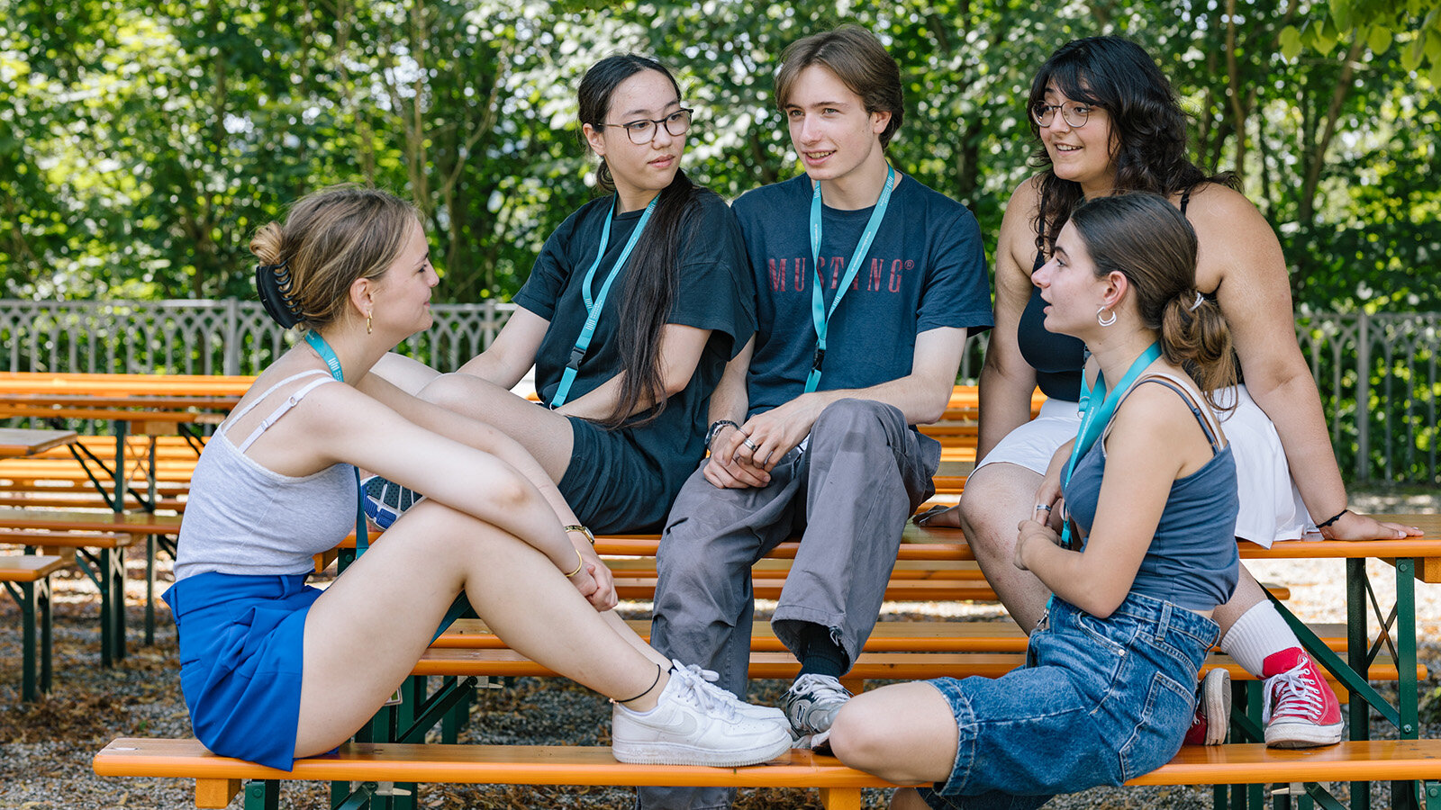 Einige junge Menschen sitzen draußen auf orangenen Bierbänken und -tischen und unterhalten sich miteinander.