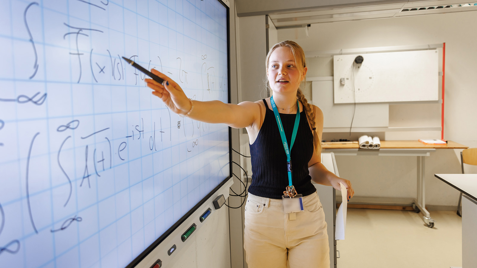 Eine junge frau zeigt mit einem Stift auf ein Smartboard, an dem komplexe mathematische Formeln zu erkennen sind.