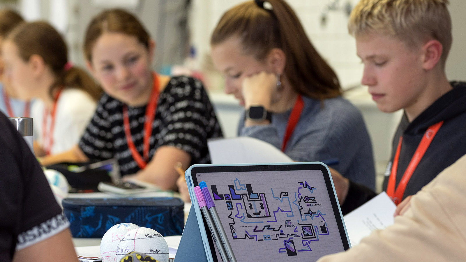 Im Vordergrund ist ein Tablet mit einem bunten Bild zu sehen, im Hintergrund sitzen junge Menschen an einem Tisch und arbeiten konzentriert.