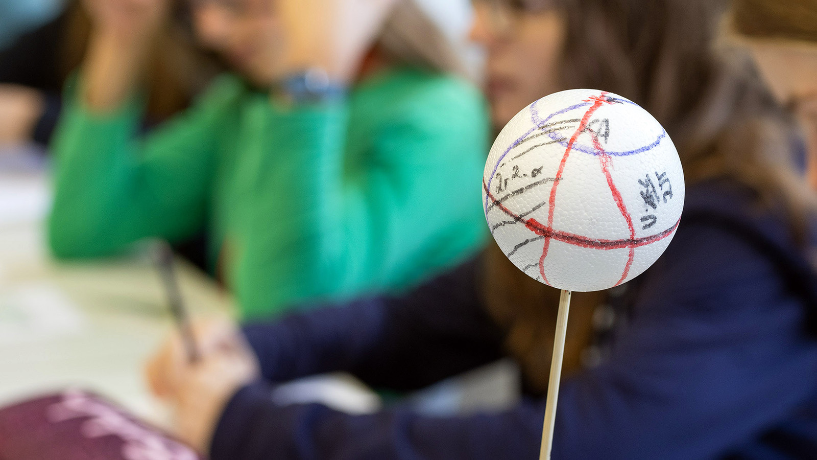 Im Vordergrund ist eine Kunststoffkugel auf einem Holzstab zu sehen, auf de rmehrere Achsen und Formeln verzeichnet wurden; im Hintergrund sind unscharf zwei Jugendliche zu sehen.