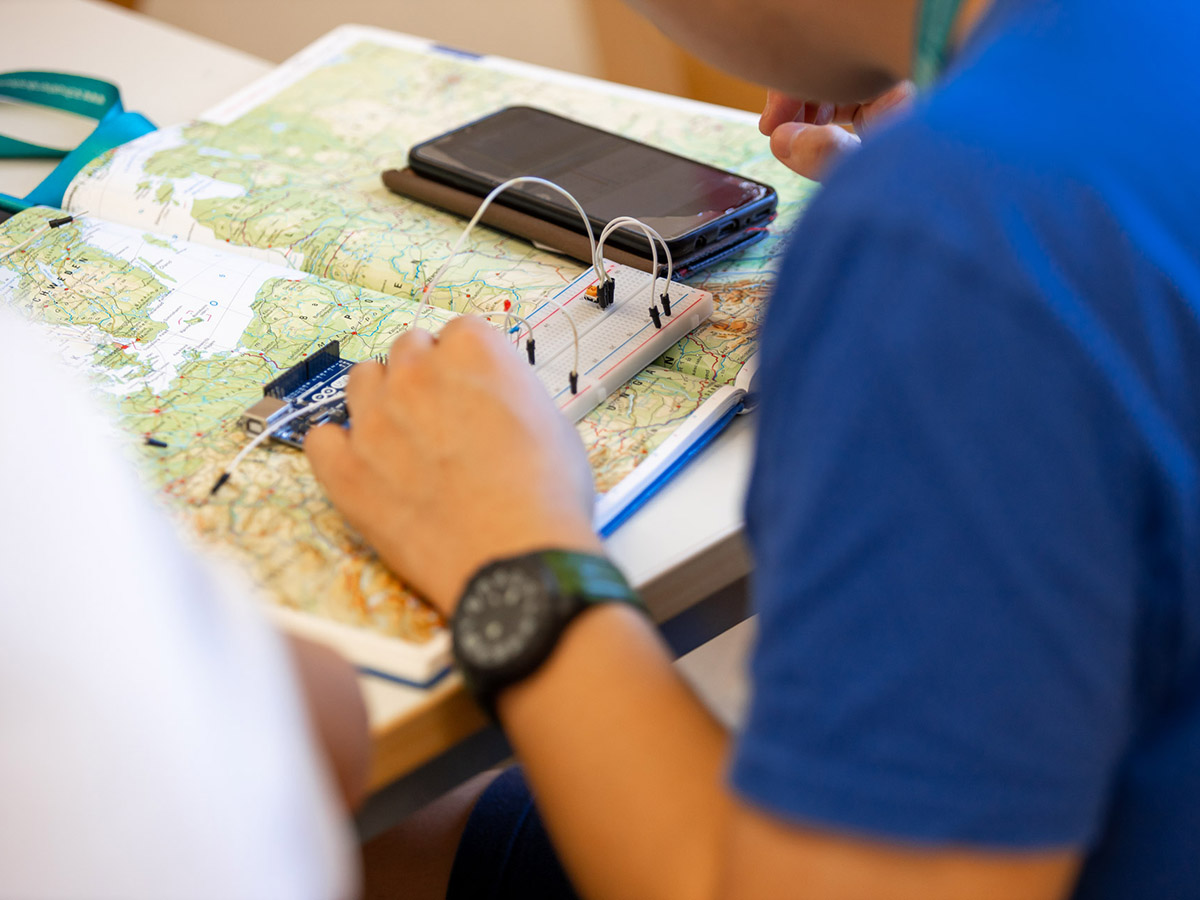 Von hinten: Eine Junge Person arbeitet an Kabeln auf eienr weißen Platine, daneben liegt ein Smartphone.