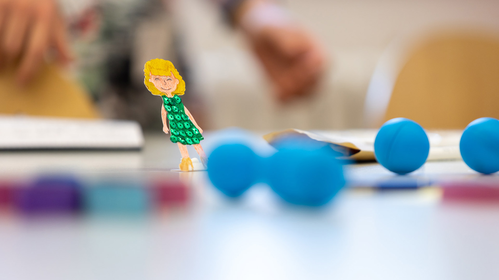 Auf einem weißen Tisch liegen mehrere blaue Kugeln. Links ist eine Papierfigur einer Frau mit grünem Kleid zu sehen.