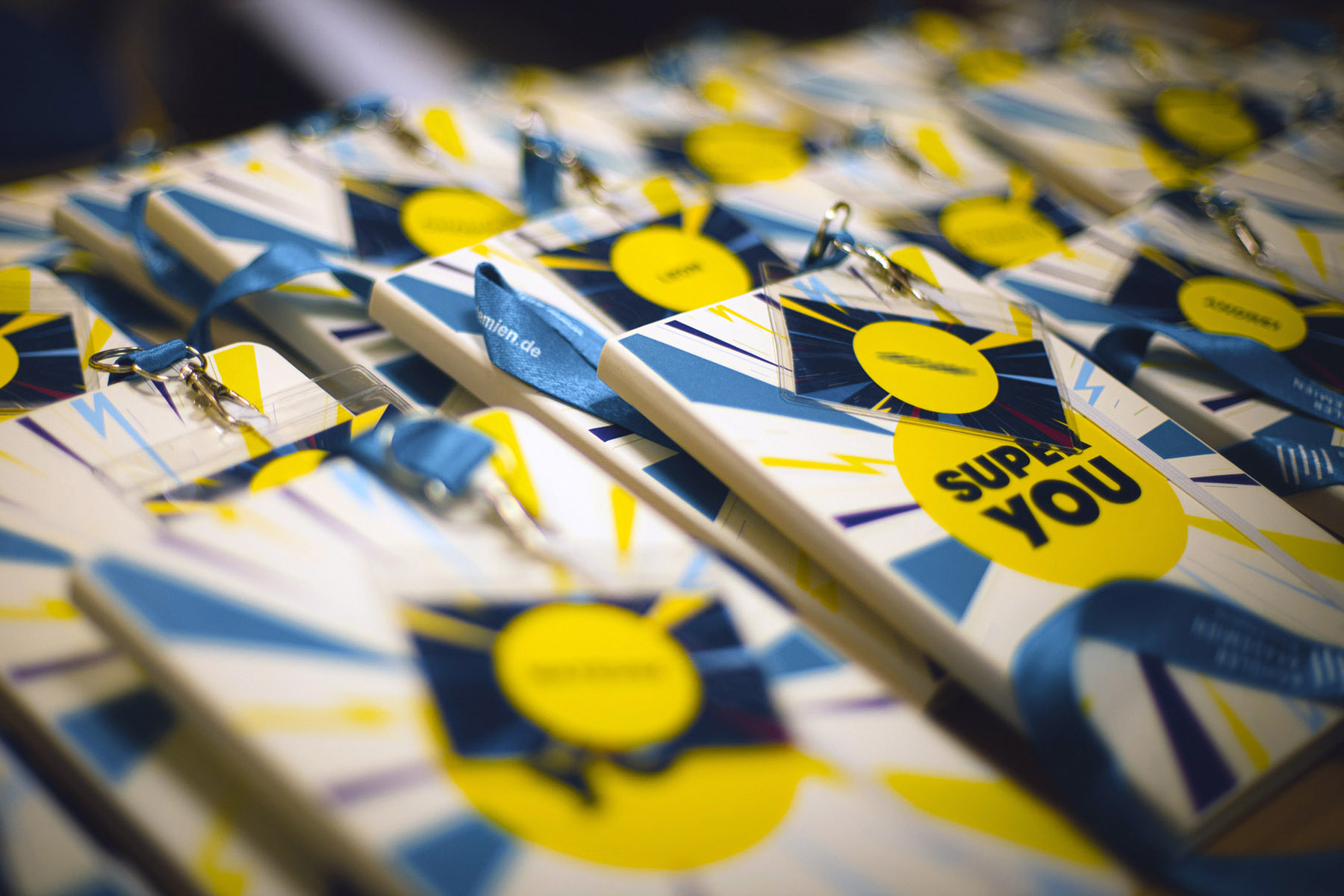 Es sind viele mit dem gelb-blau-grünen Super You Logo bedruckte Notizbücher und Namensschilder zu sehen.