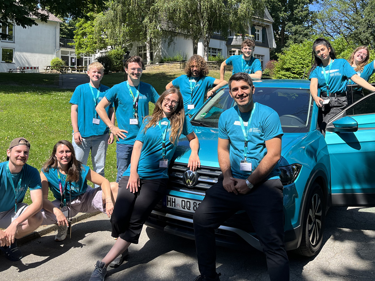 Eine Gruppe junger Menschen in petrolfarbenen T-Shirts steht rund um ein petrolfarbenes Auto, alle lächeln in die Kamera.