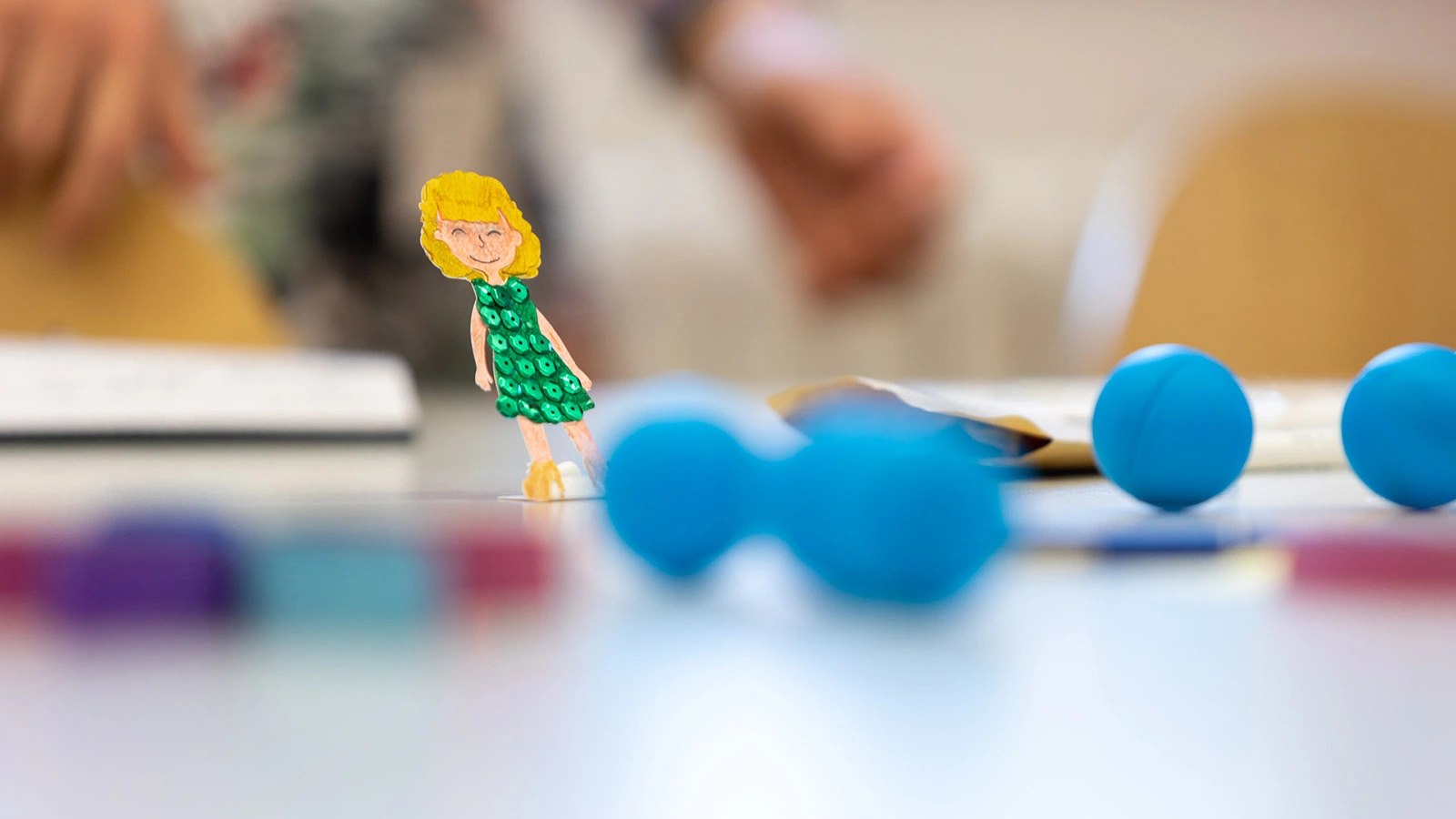Auf einem weißen Tisch liegen mehrere blaue Kugeln. Links ist eine Papierfigur einer Frau mit grünem Kleid zu sehen.